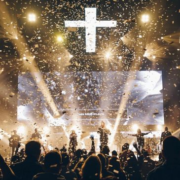 An auditorium filled with people with a band on stage and a LED cross floating in the sky.  