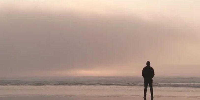 A person stands alone on a calm beach at sunset or sunrise.