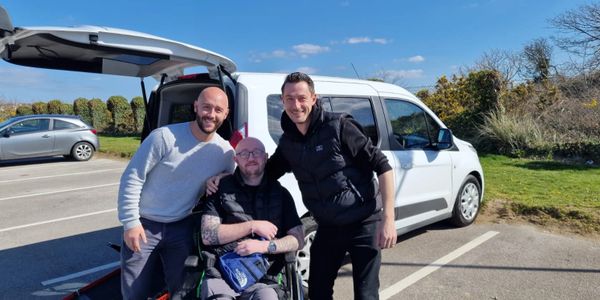 Three men smiling by a white van, one in a wheelchair, on a sunny day.