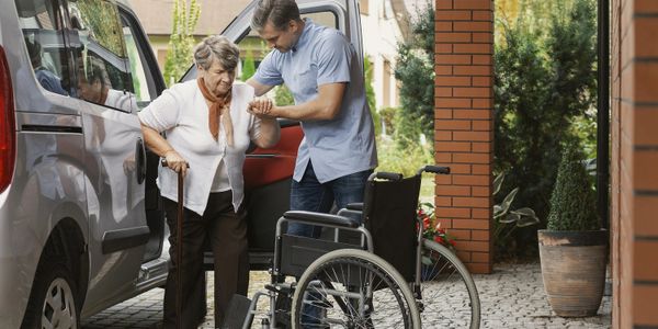 A man helps an elderly woman out of a car towards a wheelchair.