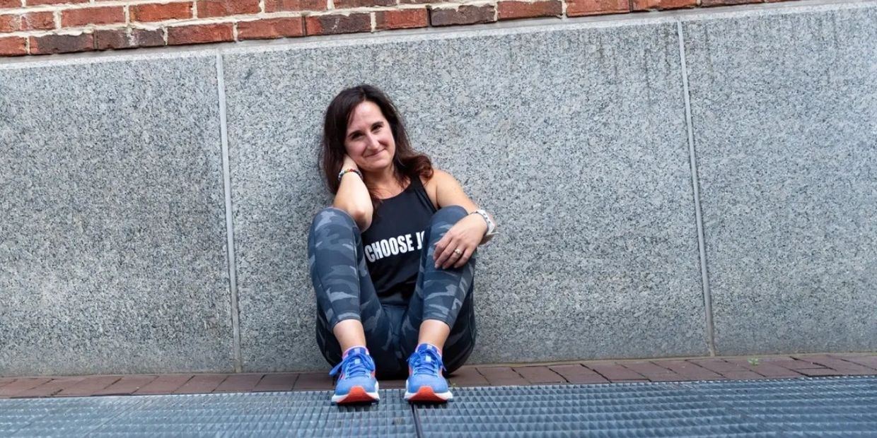 Woman in workout attire sitting against a stone wall, smiling gently.