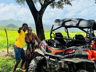 Two people smiling next to an off-road vehicle under a tree in a scenic rural area.