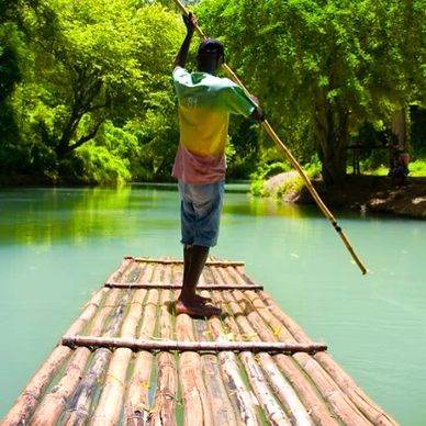 Person steering a bamboo raft on a calm river surrounded by greenery.