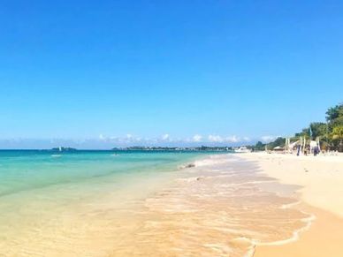A sunny beach with clear turquoise water and white sand under a bright blue sky.