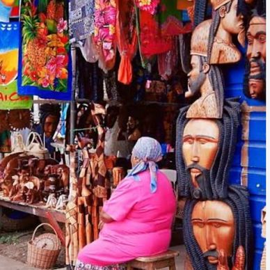 Woman in pink sitting by a vibrant market stall with wooden masks and crafts.