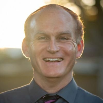 Smiling man in a dark shirt and tie with sunlight behind him.