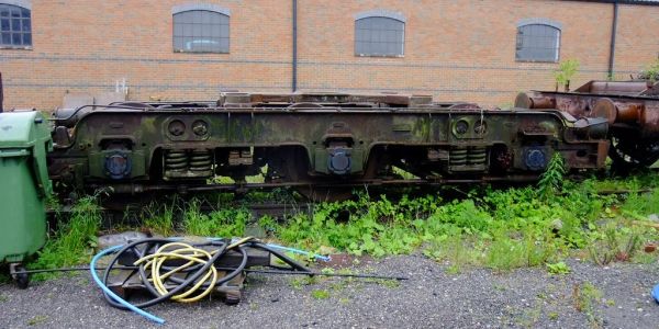 Rusty train bogie resting outdoors near a brick building and green plants.