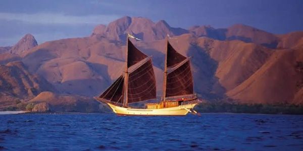 Sailboat with brown sails on blue water near mountainous shore.