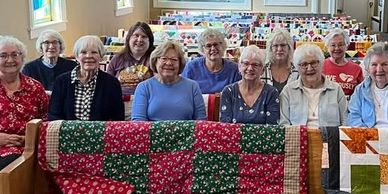 Group of elderly women smiling with colorful quilts in a cozy room.