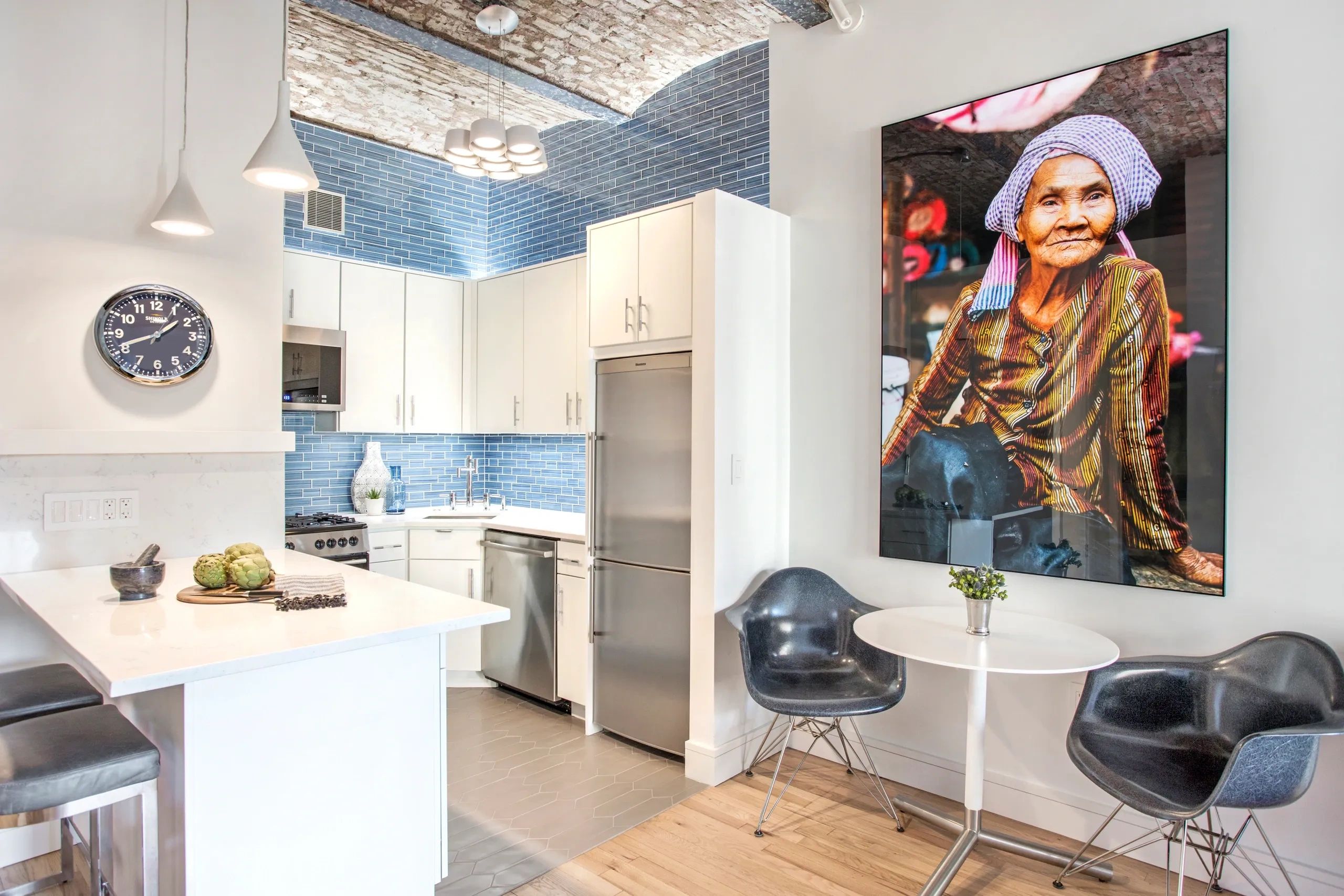 Small kitchen with blue tile, small bistro table and 2 chairs with painting of old lady on the wall. 