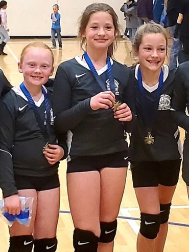 Three young volleyball players proudly display their medals in a gym.