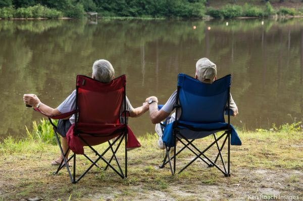 Two elderly people holding hands while sitting by a lake in camping chairs.