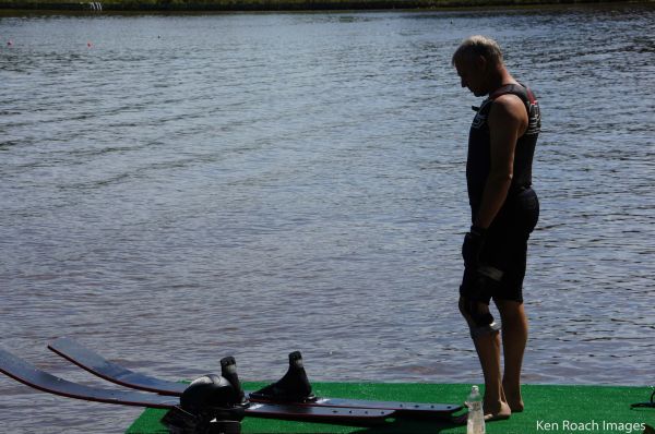 Man in wetsuit standing by water skis on a dock next to a lake.