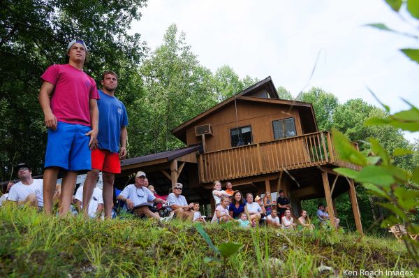 A group of people gathered outdoors near a wooden cabin in a forested area.