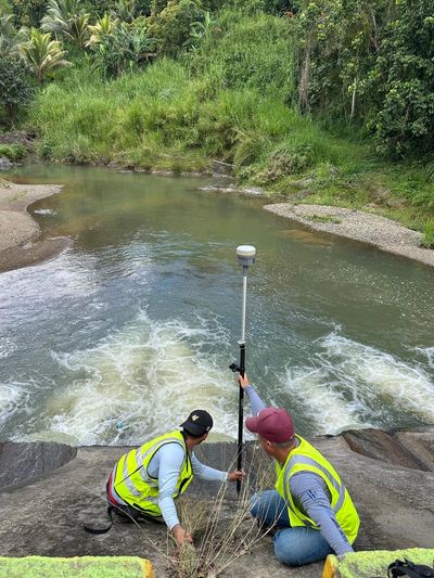 Two workers in safety vests measure water flow by a river.