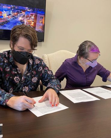 Two people wearing masks signing documents at a table.