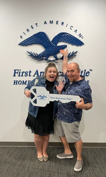 A cheerful couple holds a large key in front of First American Title sign.