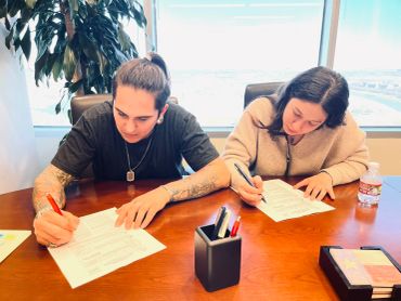 Two people focused on signing documents at a wooden table in a bright office.