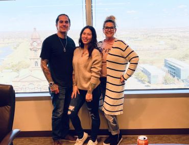 Three people smiling in front of a large window with a cityscape view.