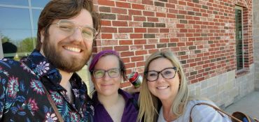 Three friends smiling and posing for a selfie outside near a brick wall.