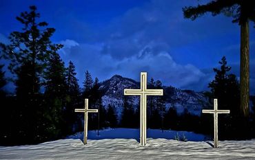 Three crosses stand illuminated in the snow against a mountain backdrop at dusk.