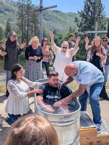 A man is being baptized outdoors in a metal tub, surrounded by people praying.