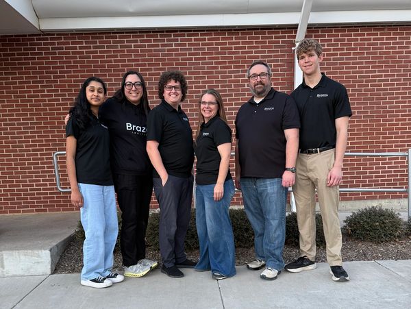 Group of six diverse people standing in front of a brick wall, smiling.