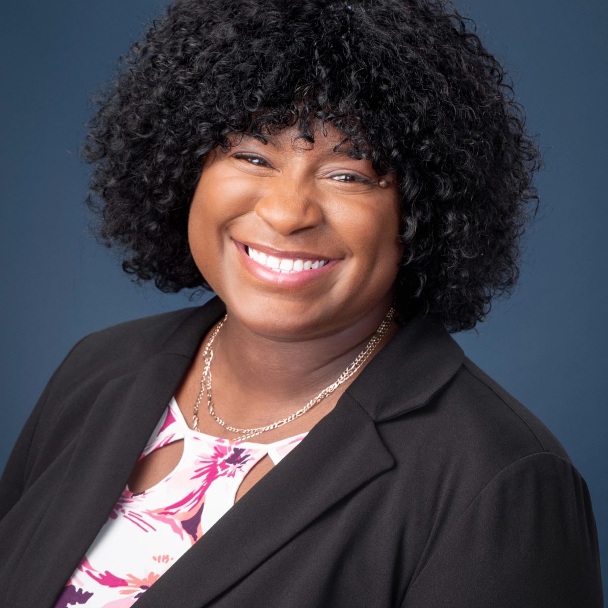 Smiling woman with curly hair in professional attire against a blue background.