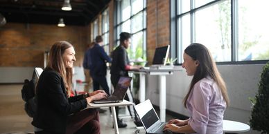 Two women working on laptops and smiling in a modern office.