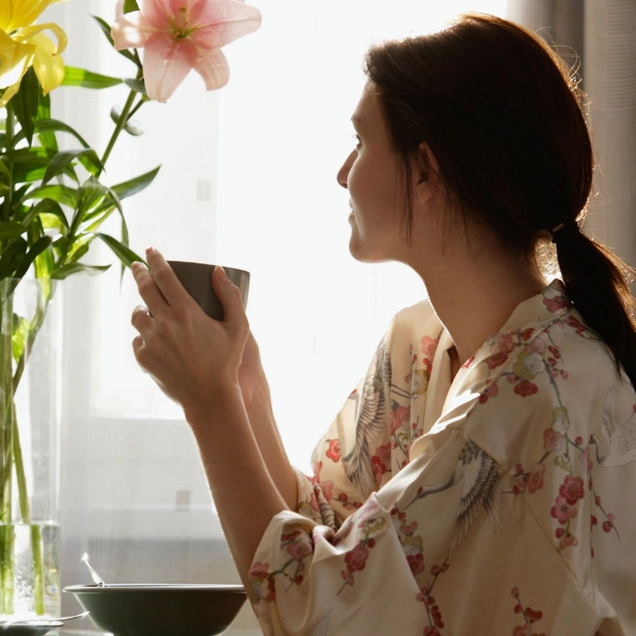 A woman holding a tea mug and looking out the window while sitting at a table with pink flowers.