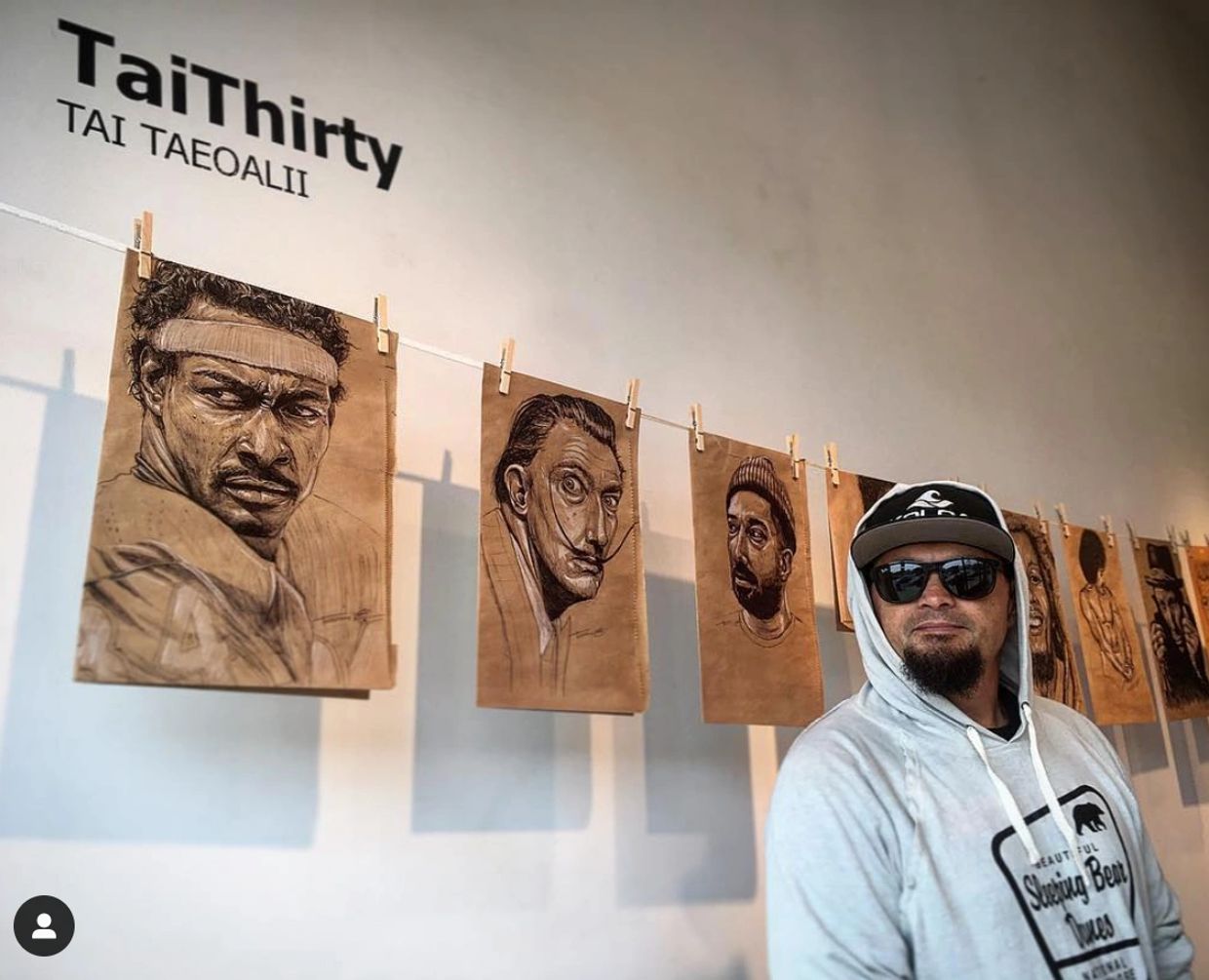 Artist posing beside his detailed portrait sketches displayed on a gallery wall.