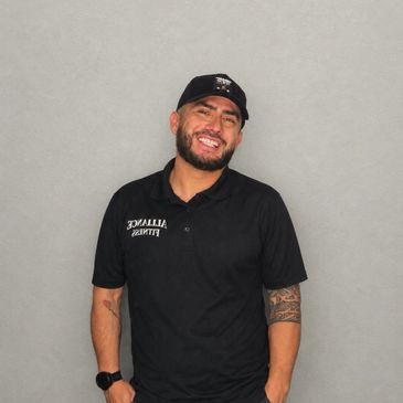 Smiling man in black polo and cap with tattoos, standing against gray background.