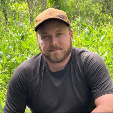 A man with a beard and cap outdoors in greenery.