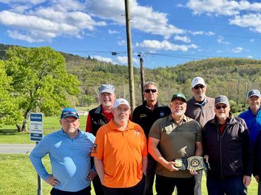 Group of men posing outdoors on a sunny day, one holding a trophy.
