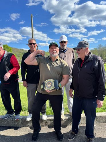 Group of men outdoors, one flexing muscles and holding a championship belt.