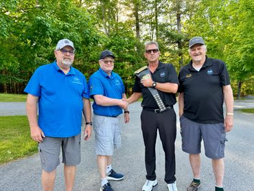 Four men outdoors, two shaking hands while holding a trophy.