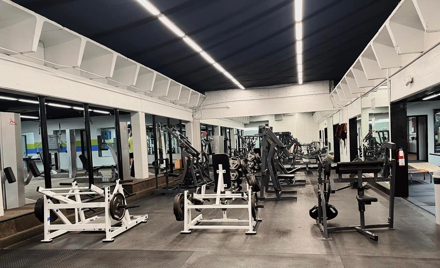 A spacious, well-lit gym with various weightlifting machines and mirrors lining the walls.