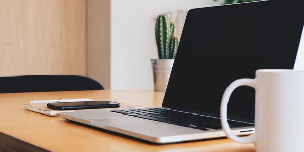 Open laptop on a wooden desk with a white coffee mug, smartphone, and small potted cactus in the background