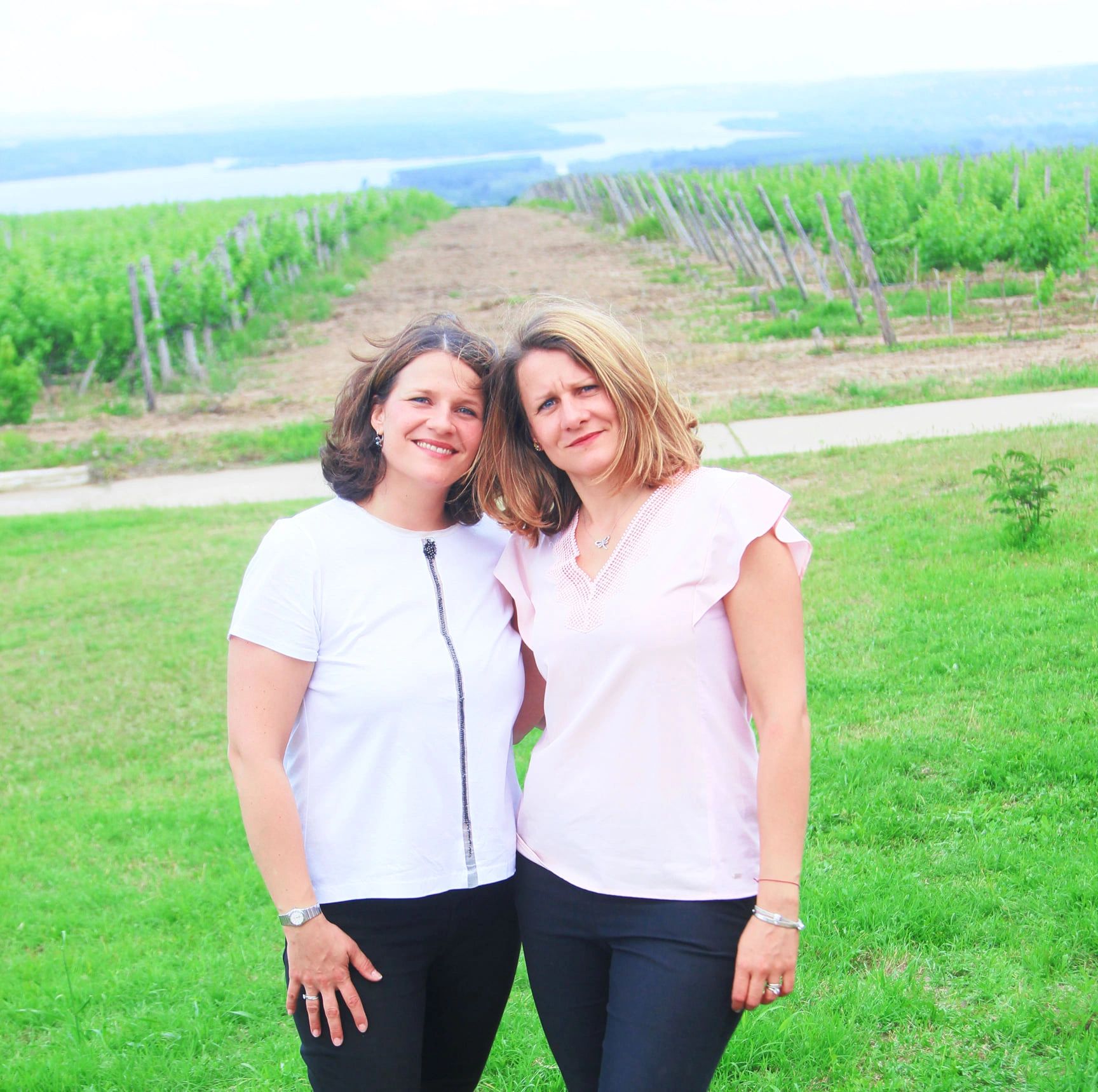 Two women posing in a vineyard with a river in the background.