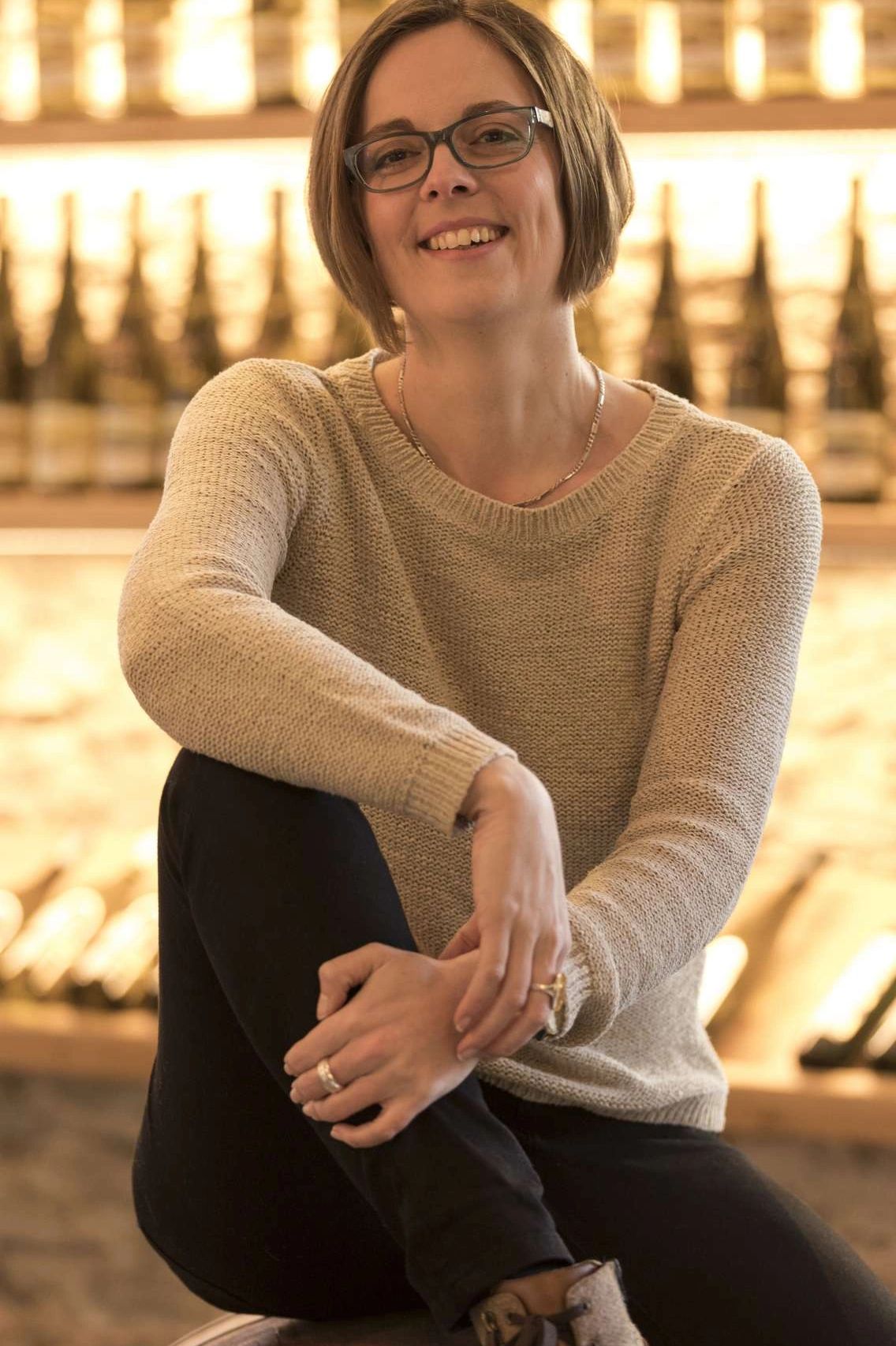 Smiling woman in glasses wearing a beige sweater and black pants.
