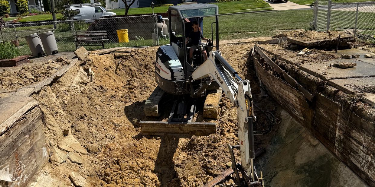 A small excavator digging in a large backyard hole with a child operating it.