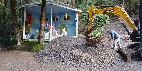 Man working with excavator on gravel pile near a small decorated building in a forest.