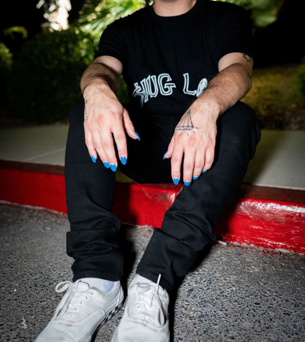 Person sitting with blue nail polish and a diamond tattoo on their hand.