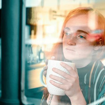 Woman with red hair holding a mug, gazing thoughtfully through a window.