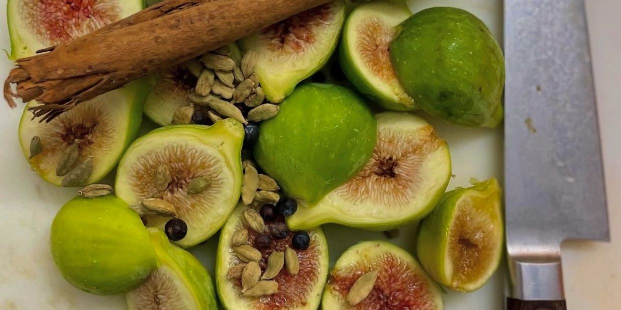 Fresh green figs, cardamom pods, cinnamon sticks, and a knife on a cutting board.