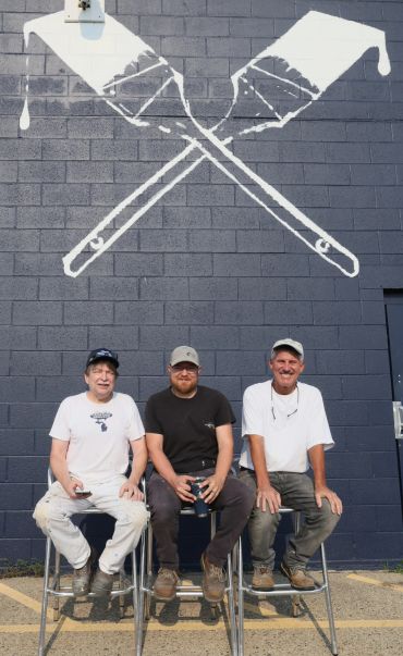 Three men sitting on stools under a wall painting of crossed paintbrushes.