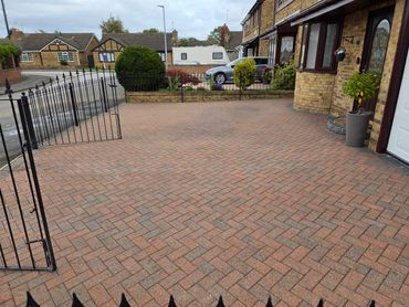 Brick-paved driveway with black metal gate and garden beds beside a brick house.