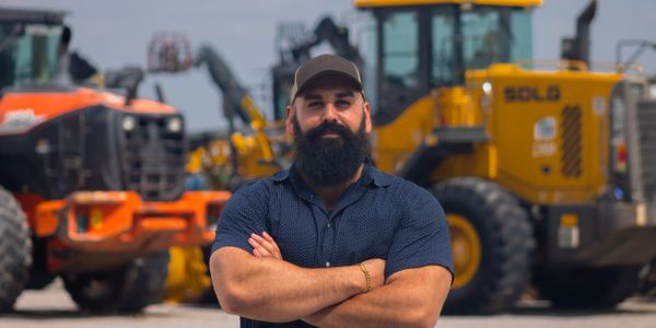 Bearded man in cap stands confidently in front of heavy construction machinery.