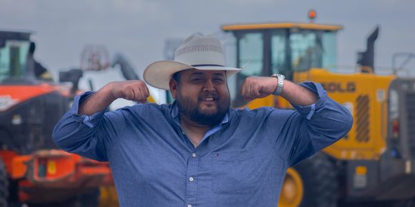 Man in cowboy hat flexing muscles near construction equipment.