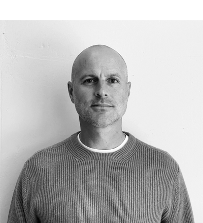 Black and white portrait of a man in a knitted sweater against a plain wall.